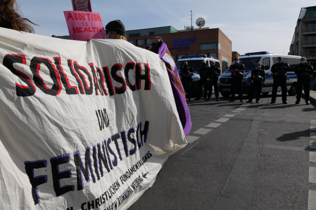 Eine Gruppe von Menschen marschiert auf einer Straße, hält eine Fahne mit der Aufschrift 'Solidarität und Feminismus' und hat parkende Fahrzeuge und Gebäude im Hintergrund unter einem klaren blauen Himmel.
