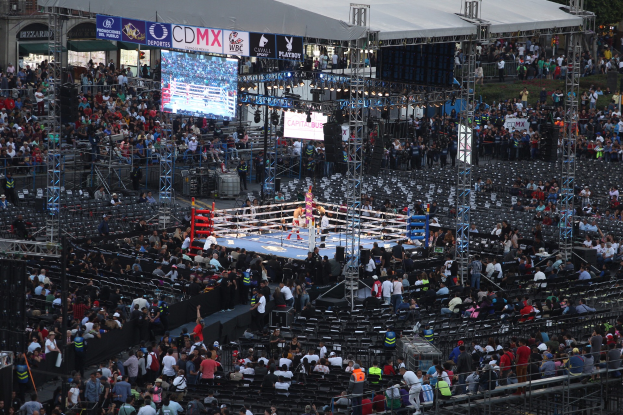 Eine große Menschenmenge in einem Stadion bei einem Boxkampf, einige stehen am Boden, andere sitzen, Metallstangen, ein Zaun, ein Display mit Text, Metallrahmen, ein Dach mit Stangen, Schilder mit Text, ein Gebäude mit Fenstern, Bäume und ein bewölkter Himmel.