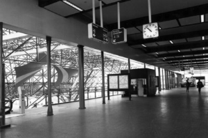 Ein Schwarz-Weiß-Foto eines Flughafen-Terminals mit Menschen, Pfählen, Brettern, einer Uhr und Deckenleuchten.