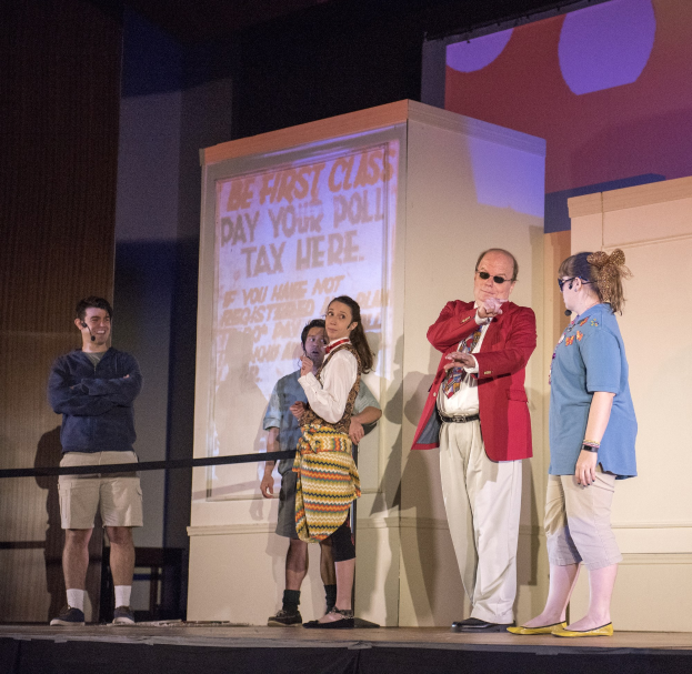 Eine Gruppe von Menschen auf einer Bühne mit einem Mann, der ein Mikrofon hält, ein Display mit der Aufschrift "Der erste Klasse-Tag Deine Steuer hier", ein Zaun, eine Wand und Deckenleuchten.