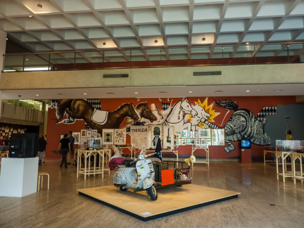 Ausstellungshalle im Nationalen Kunstmuseum mit Tischen, Stühlen, einem Motorroller, einem Fernseher, stehenden Besuchern, Foto Rahmen, einer Wandmalerei und Deckenlampen.