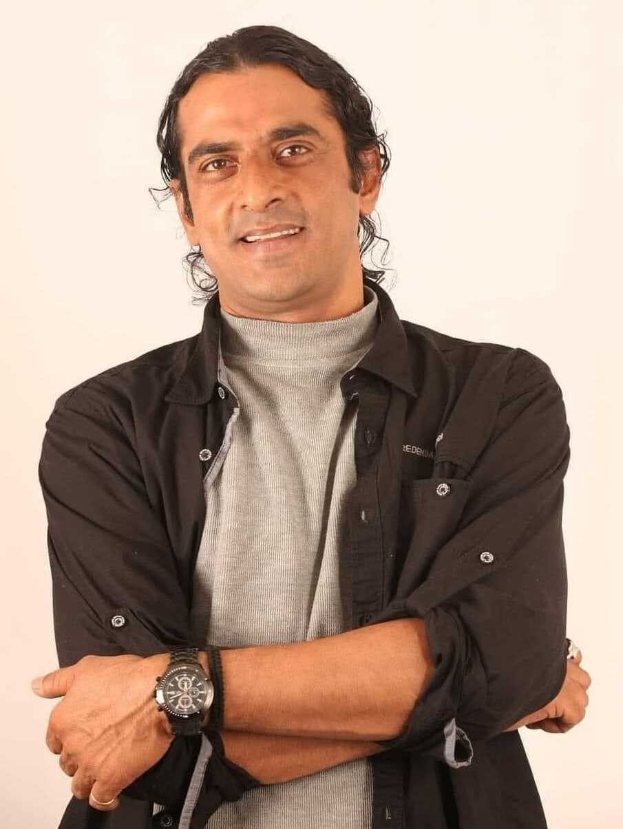 Ein Mann in einem schwarzen und aschfarbenen Kleid, der eine Uhr trägt, steht mit überkreuzten Armen vor einem weißen Hintergrund und sieht nachdenklich aus.