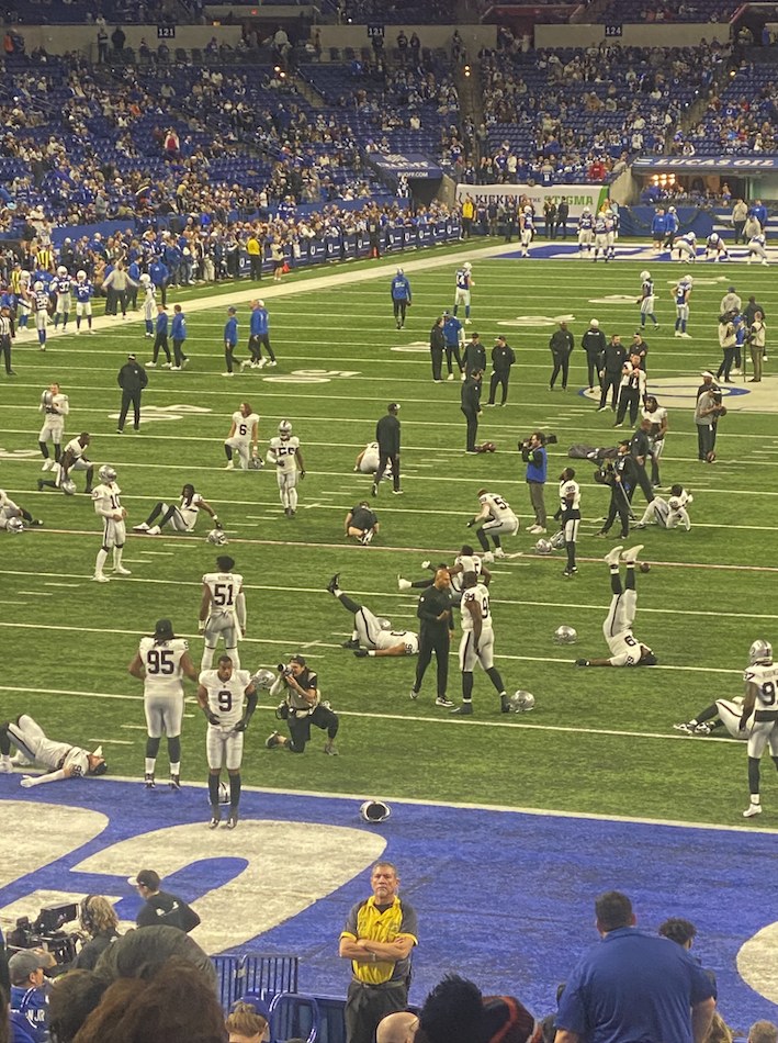 Ein Fußballspiel in einem Stadion mit Spielern auf dem Feld und Zuschauern im Hintergrund, einschließlich einer Person, die eine Kamera für die Live-Übertragung bedient.