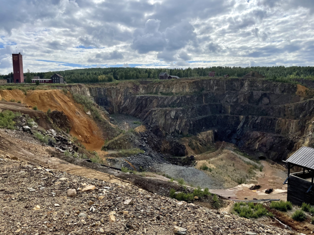 Großer Tagebau auf einem Schotterfeld umgeben von Bäumen, Gebäuden und einem Schuppen, mit bewölktem Himmel und Boden voller Steine und Pflanzen.