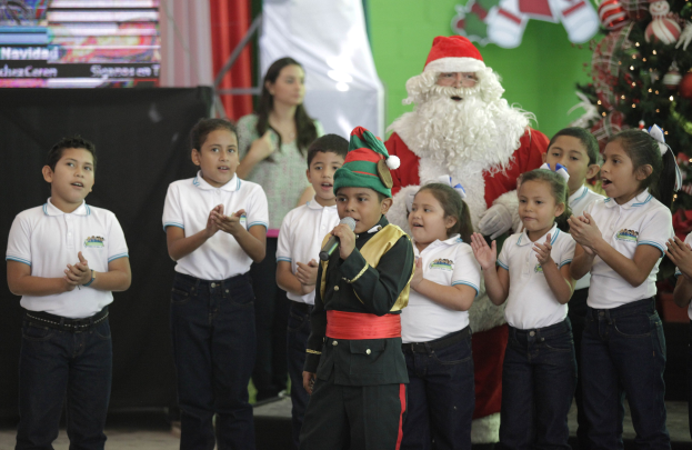 Kinder klatschen neben Santa Claus vor einem geschmückten Weihnachtsbaum, wobei ein Kind ein Mikrofon hält und eine Frau im Hintergrund steht.