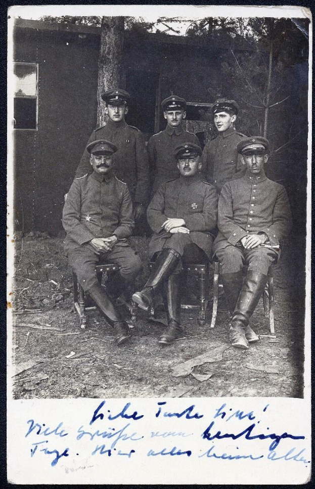 Schwarzes und weißes Foto einer Gruppe deutscher Soldaten in Uniformen des Ersten Weltkriegs, die auf Stühlen vor einem Haus mit Bäumen im Hintergrund sitzen.