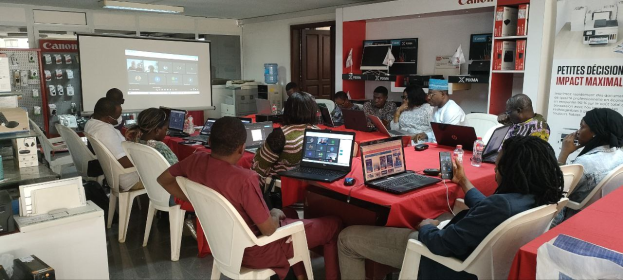 Gruppe von Menschen um einen Tisch mit Laptops und anderen Gegenständen während eines digitalen Marketing-Workshops, mit einem Banner, einem Display-Bildschirm und einer Tür im Hintergrund.