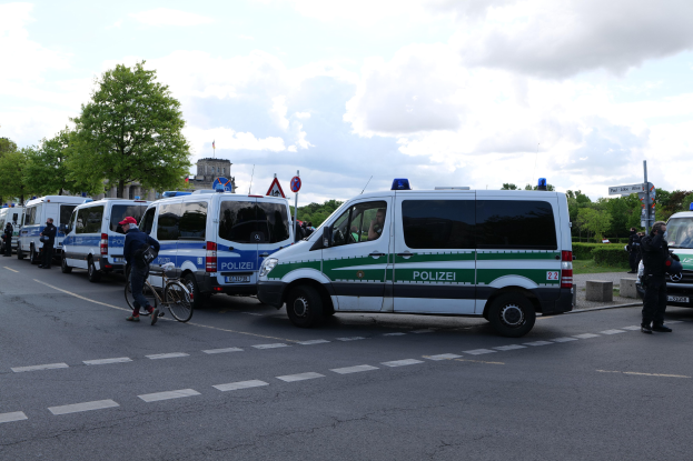Eine Gruppe von Polizeiwagen, die entlang einer Straße geparkt sind, mit einem Radfahrer im Vordergrund, Bäumen, Masten, Schildern, Gebäuden und einem bewölktem Himmel im Hintergrund.