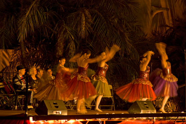 Menschen tanzen auf einer Bühne mit Scheinwerfern und Bäumen im Hintergrund, während andere auf Stühlen im Dunkeln sitzen.