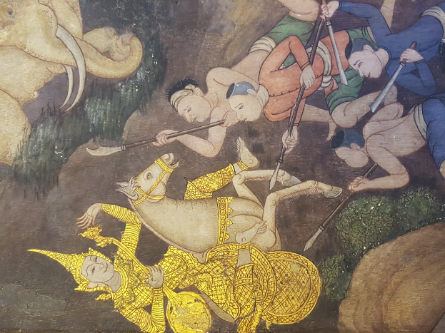 Ein farbenfrohes und detailreiches Wandgemälde auf einer buddhistischen Tempelwand in Thailand zeigt einen zentralen Elefanten mit erhobenem Rüssel umgeben von Menschen in traditioneller thailändischer Kleidung, die in Aktivitäten wie Musik und Gesang vertieft sind.