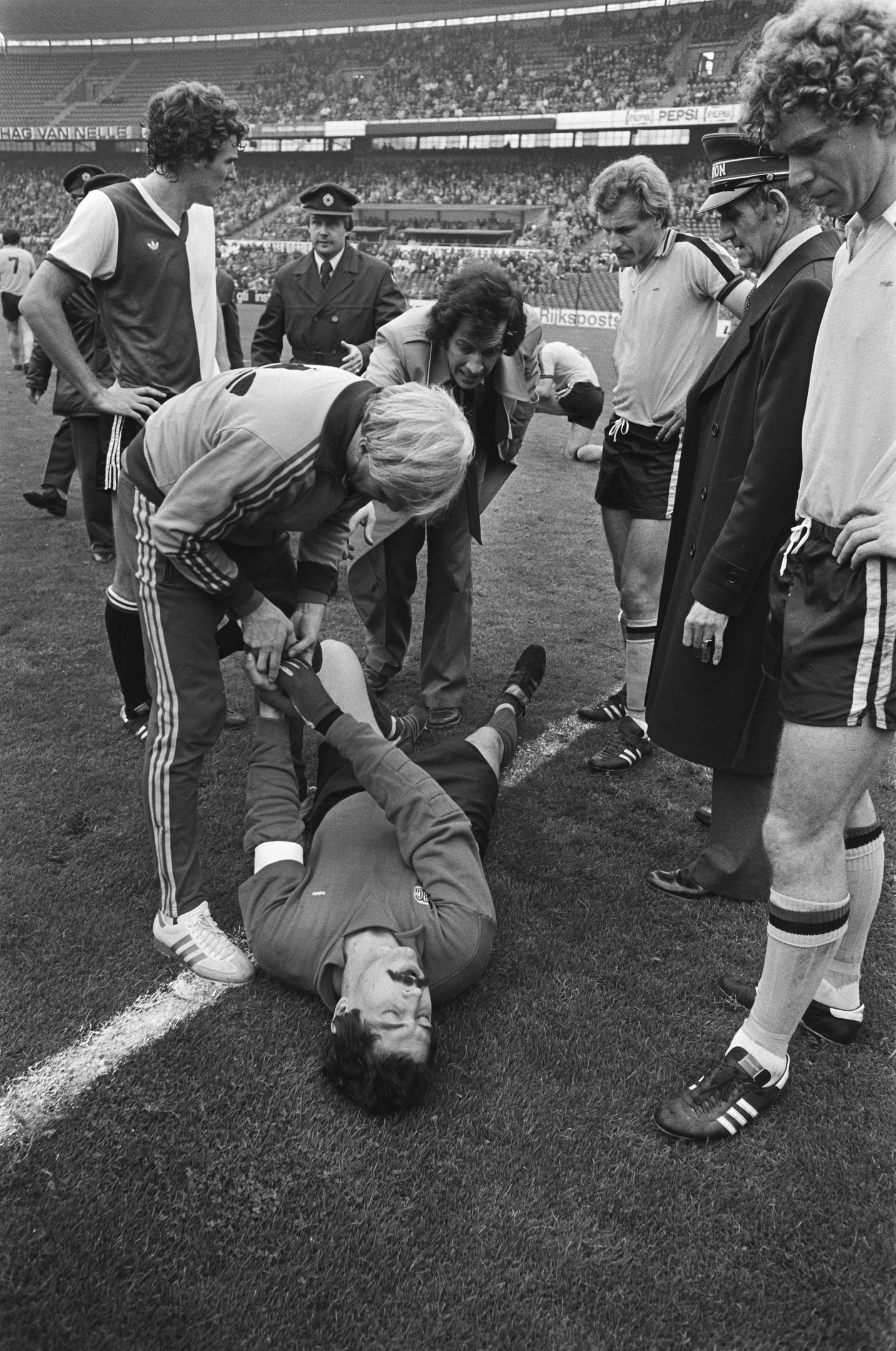 Schwarzes und weißes Bild eines Fußballspielers, der auf dem Boden liegt und von einer Gruppe Männer umgeben ist, während eine Menge zusieht.