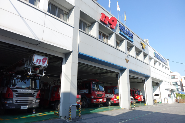 Feuerwache mit mehreren Feuerwehrautos vor der Tür, mit Fenstern, Säulen und Namensschildern, mit Fahnen im Vordergrund und anderen Gebäuden gegen einen klaren blauen Himmel.