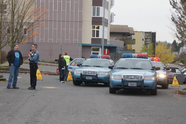Vier Autos auf einer Straße, vier Menschen in der Nähe, Gebäude mit Fenstern im Hintergrund, Bäume und ein Warndreieck.