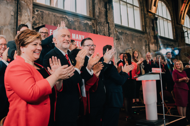 Eine Gruppe von Menschen, die vor einem Publikum stehen und klatschen, mit einem Podium, einem Mikrofon und einer Tafel mit Text auf der rechten Seite und Stühlen, einer Fahne, einer Wand, Fenstern und Lichtern im Hintergrund.
