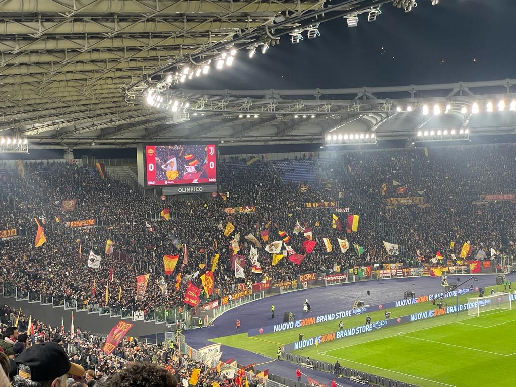 Eine große Menschenmenge in einem Stadion schwenkt Fahnen und Banner, mit Menschen in Mützen auf den Rängen, einem Bildschirm im Hintergrund und beleuchteter Decke, was die Vorfreude auf ein Fußballspiel vermuten lässt.