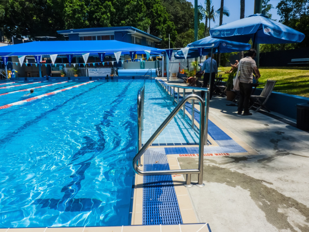 Großer Außenpool mit schwimmenden Menschen, umgeben von Geländern, Sonnenschirmen, Stühlen, Tischen, Pflanzen, Gras, Bäumen und einem Gebäude unter einem klaren blauen Himmel.