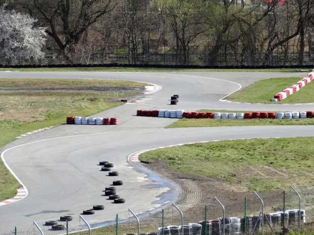 Rennstrecke mit zahlreichen Reifen, die an ihren Rändern angeordnet sind, von Gras und Maschendrahtzaun umgeben, mit Bäumen im Hintergrund.