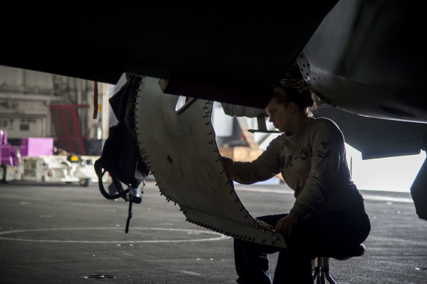 Eine Frau in Arbeitskleidung sitzt auf einem Hocker vor einem Flugzeug und hält ein Objekt in der Hand, mit Fahrzeugen und Geräten im Hintergrund.