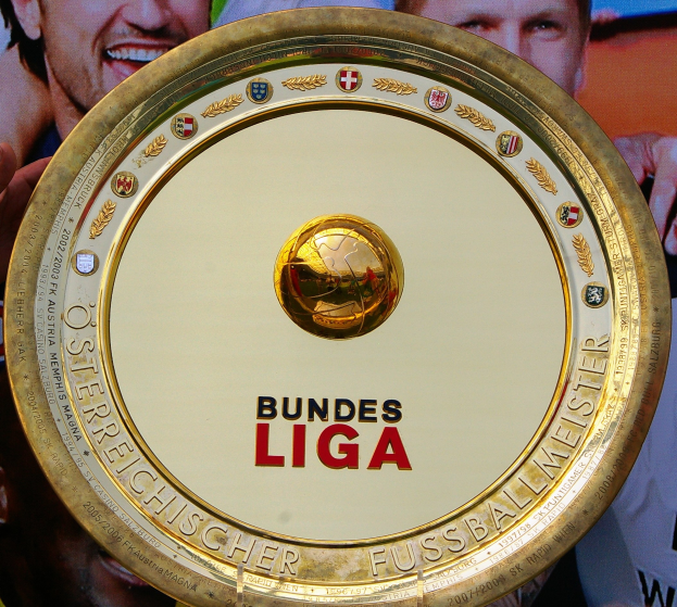 Ein Mann mit einem goldenen Schild mit 'Bundesliga' darauf, im Hintergrund eine Fahne mit drei Personen.