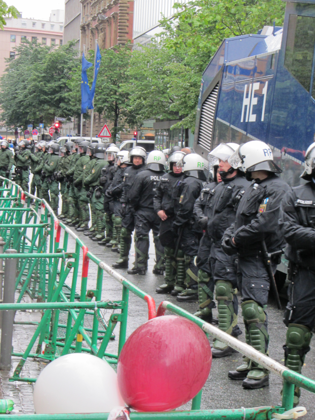Eine Gruppe von Polizisten in Helmen und mit Waffen steht vor einer Barriere auf einer Straße mit Geländern und Luftballons, mit Bäumen, Gebäuden mit Fenstern, Flaggen mit Stangen und einem klaren blauen Himmel im Hintergrund.