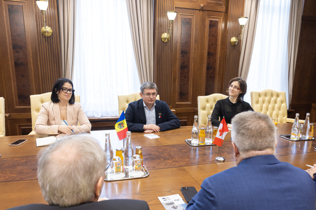 Eine Gruppe von Menschen, die um einen Holztisch in einem formellen Setting sitzen und sich mit EU-bezogenen Materialien unterhalten, umgeben von Flaggen, Flaschen, Gläsern, Papieren und Mobiltelefonen.