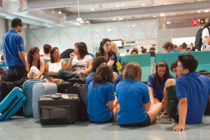 Ein belebter Flughafen mit vielen Wartenden auf Flüge, Gepäck auf der linken Seite und Glasdoors mit Richtungsschildern im Hintergrund.