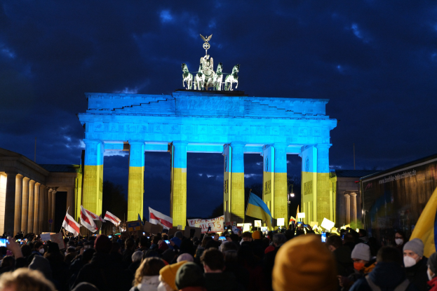 Eine Menschenmenge mit Fahnen und Plakaten steht vor dem Brandenburger Tor in Berlin, Deutschland, mit einer Fahne auf der rechten Seite und den Statuen und Säulen des Tors unter einem bewölkten Himmel.