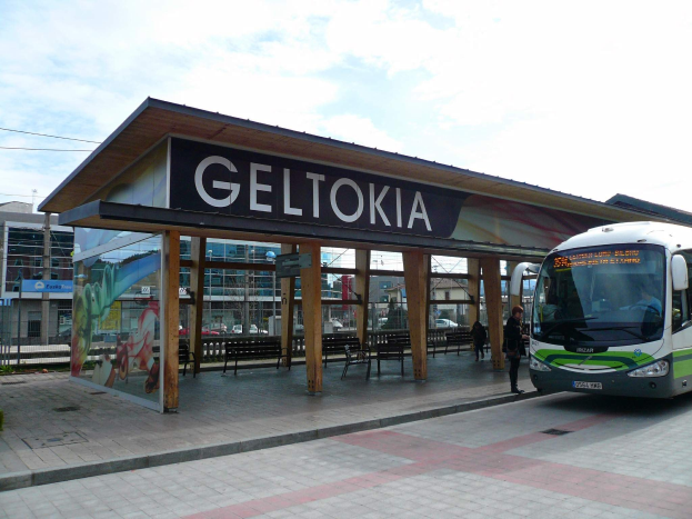 Ein Bus hält an einer Bushaltestelle mit einem "Geltokia"-Schild, Menschen in der Nähe und Gebäude, Bäume und einen klaren Himmel im Hintergrund.