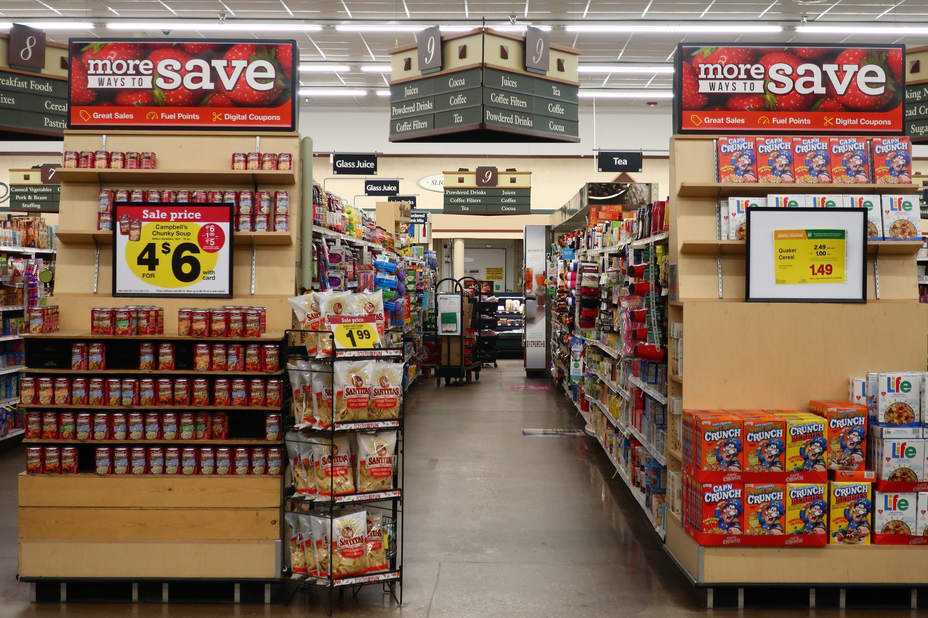 Ein Innenraum eines Lebensmittelladens mit verschiedenen Lebensmitteln auf Regalen, Regalen und dem Boden, das informative Schilder, Deckenbeleuchtung und eine Person im Hintergrund zeigt.