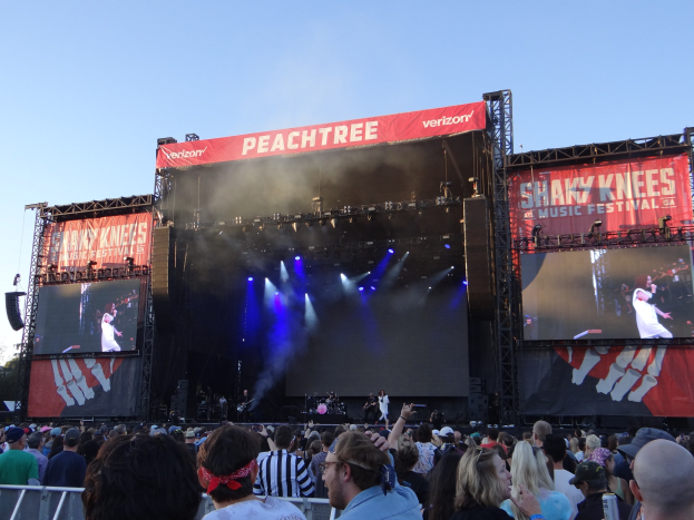 Eine große Menschenmenge steht vor einer hell erleuchteten Bühne auf dem Shaky Knees Music Festival, auf der Musiker unter Bannern mit Text spielen, mit einem Geländer im Vordergrund und einem klaren blauen Himmel im Hintergrund.