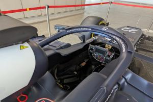 Cockpit eines Formel-E-Autos in einer Ausstellung mit einem prominenten Lenkrad, einer Wand im Hintergrund und dekorativen Bändern an den umliegenden Pfosten.