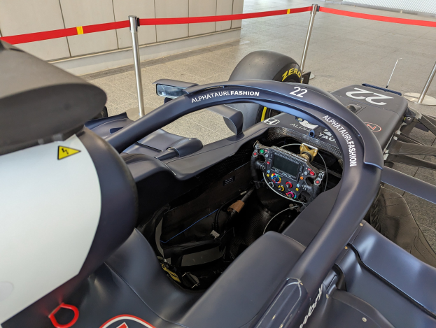 Cockpit eines Formel-E-Autos in einer Ausstellung mit einem prominenten Lenkrad, einer Wand im Hintergrund und dekorativen Bändern an den umliegenden Pfosten.