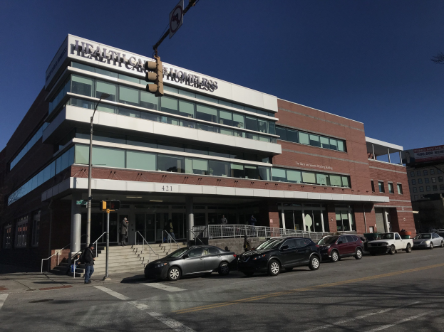 Großes Gebäude als das New York City Health Care Obdachlosen-Zentrum mit geparkten Autos, Fußgängern, Straßenmöbeln und Bäumen unter einem klaren Himmel.