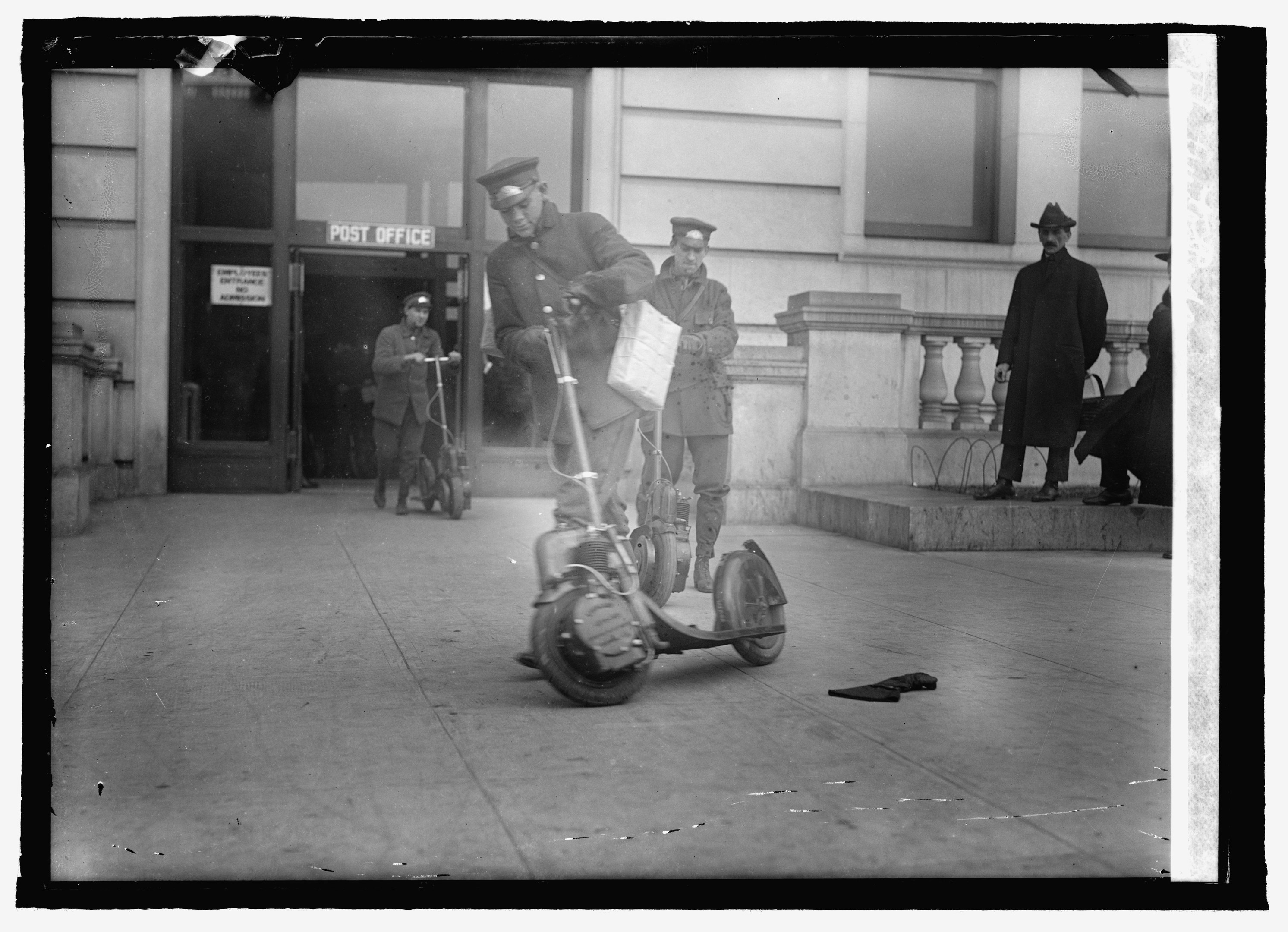 Schwarz-weißes Foto eines Mannes mit Mütze auf einem Elektroroller auf einem Gehweg, umgeben von anderen Menschen, darunter ein Radfahrer, und einem Gebäude mit einer Tür und einem Schild im Hintergrund.