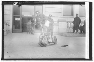Schwarz-weißes Foto eines Mannes mit Mütze auf einem Elektroroller auf einem Gehweg, umgeben von anderen Menschen, darunter ein Radfahrer, und einem Gebäude mit einer Tür und einem Schild im Hintergrund.