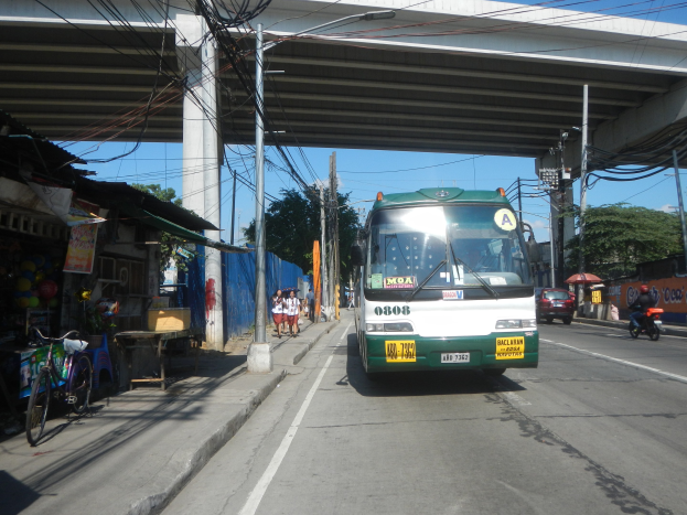 Ein grüner und weißer Bus fährt eine Straße entlang, die von Strommasten und -leitungen gesäumt ist, vorbei an Geschäften und Ständen, mit Menschen auf dem Gehweg und Fahrrädern in der Nähe, während Bäume und ein klarer blauer Himmel im Hintergrund zu sehen sind.