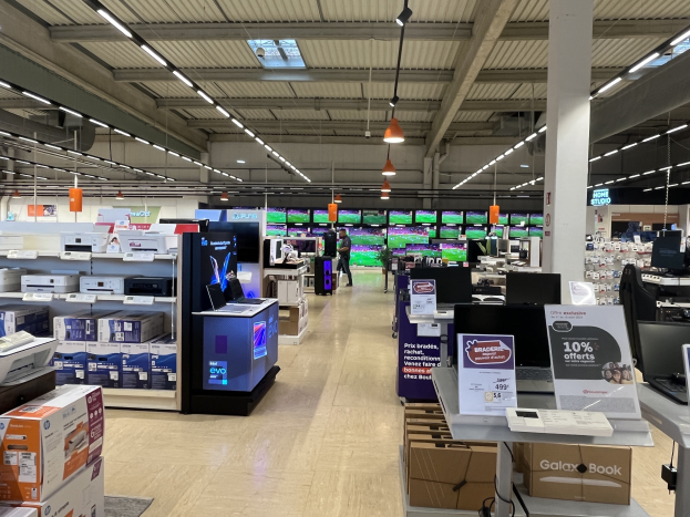 Ein großer Raum in einem Walmart-Laden voller Elektronik, die in Regalen angeordnet ist, einschließlich Computer, Drucker und Kartons, mit Menschen auf dem Boden, einer Säule, Schildern, Deckenleuchten und Deckenventilatoren.