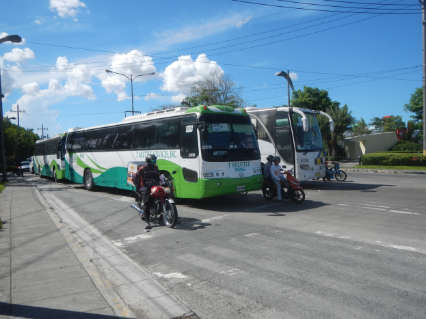Ein grüner und weißer Shuttlebus steht am Straßenrand mit Motorradfahrern davor, ein grasbewachsener Fußweg links daneben und im Hintergrund Gebäude, Bäume und Laternen unter einem klaren blauen Himmel.