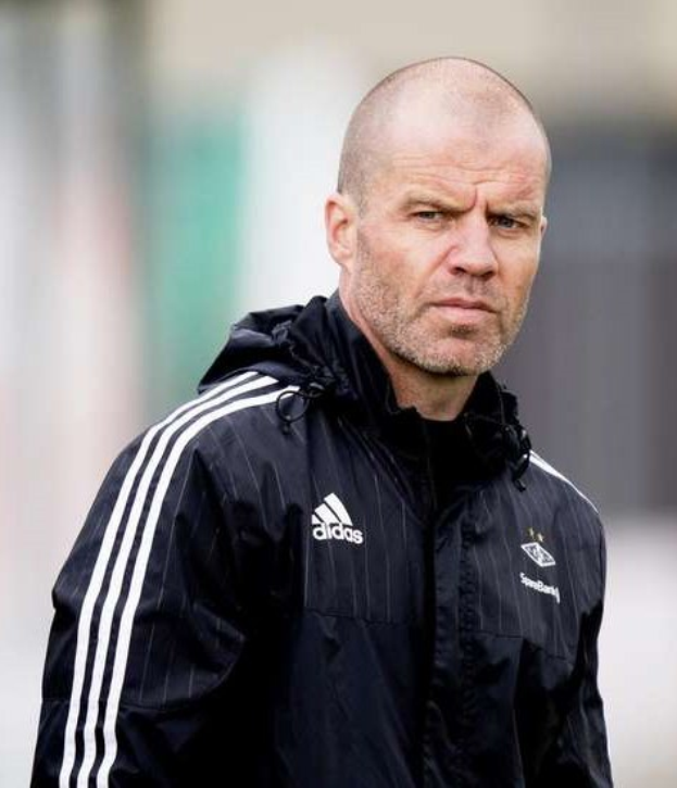 Ein kahlköpfiger Mann in einer schwarzen Jacke steht auf einem Fußballfeld und trägt eine marineblaue Jacke mit weißen Streifen und hat die Hände vor der Brust verschränkt.