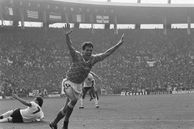 Ein Schwarz-Weiß-Foto eines Fussballspielers, der nach einem Tor feiert, umgeben von Menschen auf dem Boden, mit Schildern, Fahnen und Pfosten im Hintergrund unter einem klaren Himmel.