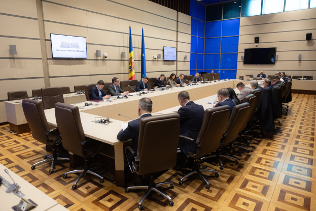 Große Gruppe von Menschen an einem langen Tisch mit Mikrofonen und Papieren in einer formellen Besprechungssituation, mit EU-Flaggen, Fernsehern und Deckenleuchten im Hintergrund.
