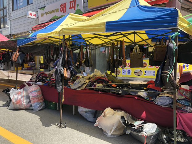 Ein Straßenmarkt mit Zelten, die Taschen, Huts und andere Gegenstände präsentieren, vor Häusern mit Fenstern und Schildern im Hintergrund.