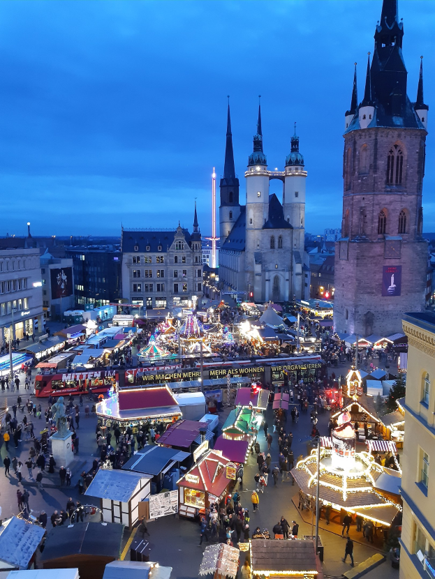 Ein belebter Weihnachtsmarkt in Köln, Deutschland, bei Nacht mit Menschen, die von Stand- und Gebäudelichtern beleuchtet werden, unter einem bewölkten Himmel.