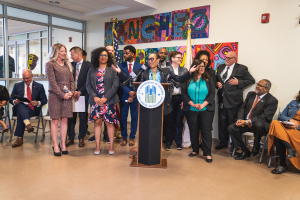 Eine Gruppe von Menschen steht und sitzt in einem Raum mit einem Podium, einem Mikrofon und einer Tafel mit der Aufschrift "Sancho City Councilman Joins City Council", mit Flaggen, einer Tafel, einem Feuerlöscher und Deckenleuchten an der Rückwand.