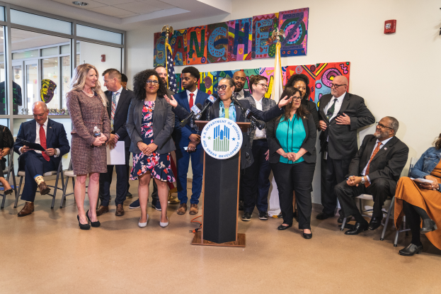 Eine Gruppe von Menschen steht und sitzt in einem Raum mit einem Podium, einem Mikrofon und einer Tafel mit der Aufschrift "Sancho City Councilman Joins City Council", mit Flaggen, einer Tafel, einem Feuerlöscher und Deckenleuchten an der Rückwand.