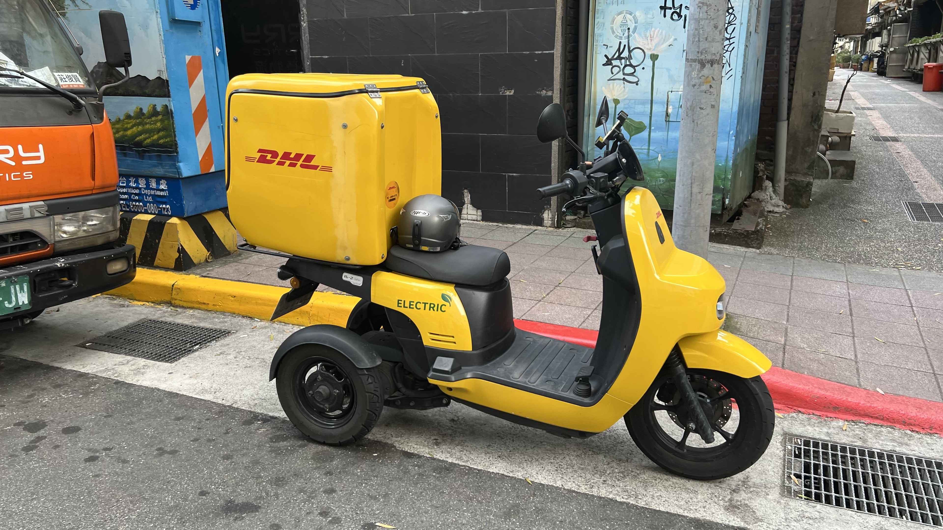 Ein gelbes Elektro-Scooter mit Helm steht auf dem Bürgersteig neben einem Lastwagen geparkt, mit Gebäuden im Hintergrund und ein paar Menschen, die auf dem Gehweg rechts gehen.