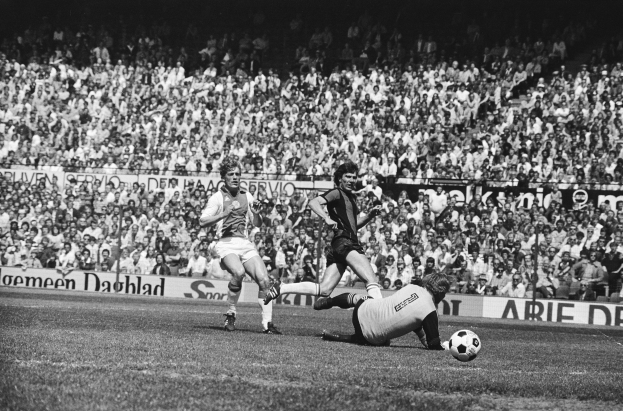 Schwarzes und weißes Foto von Männern, die Fußball auf einem Feld spielen, mit Zuschauern im Hintergrund und einer 'Bundesliga 1978-79 Ajax 1-1 Hannover 96'-Fahne.