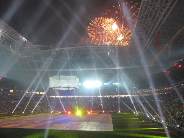Olympiastadion in London bei Nacht, erhellt von hellen Lichtern, mit einer großen Menschenmenge, saftig grünem Rasen und einem spektakulären Feuerwerk, das den Himmel erleuchtet.