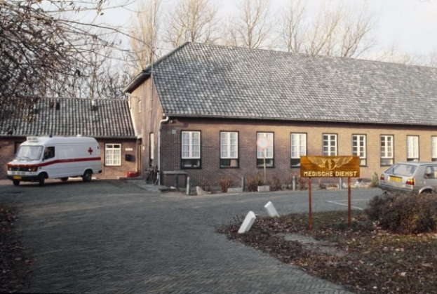 Ambulanz vor einem Backsteingebäude mit Fenstern, einem Schild, Pflanzen, Bäumen und einem klaren blauen Himmel geparkt