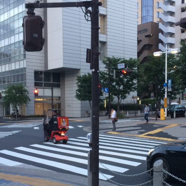 Ein Scooter auf der Straße mit Personen im Hintergrund, eine Ampel im Vordergrund und Gebäude mit Bäumen dahinter.